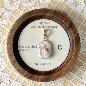May include: A natural pearl pendant with hand-applied gold leaf, set in an 18K gold clip-on charm clasp. The pendant is displayed on a white surface within a wooden frame, with the text "Natural pearl" and "Hand-applied gold leaf".
