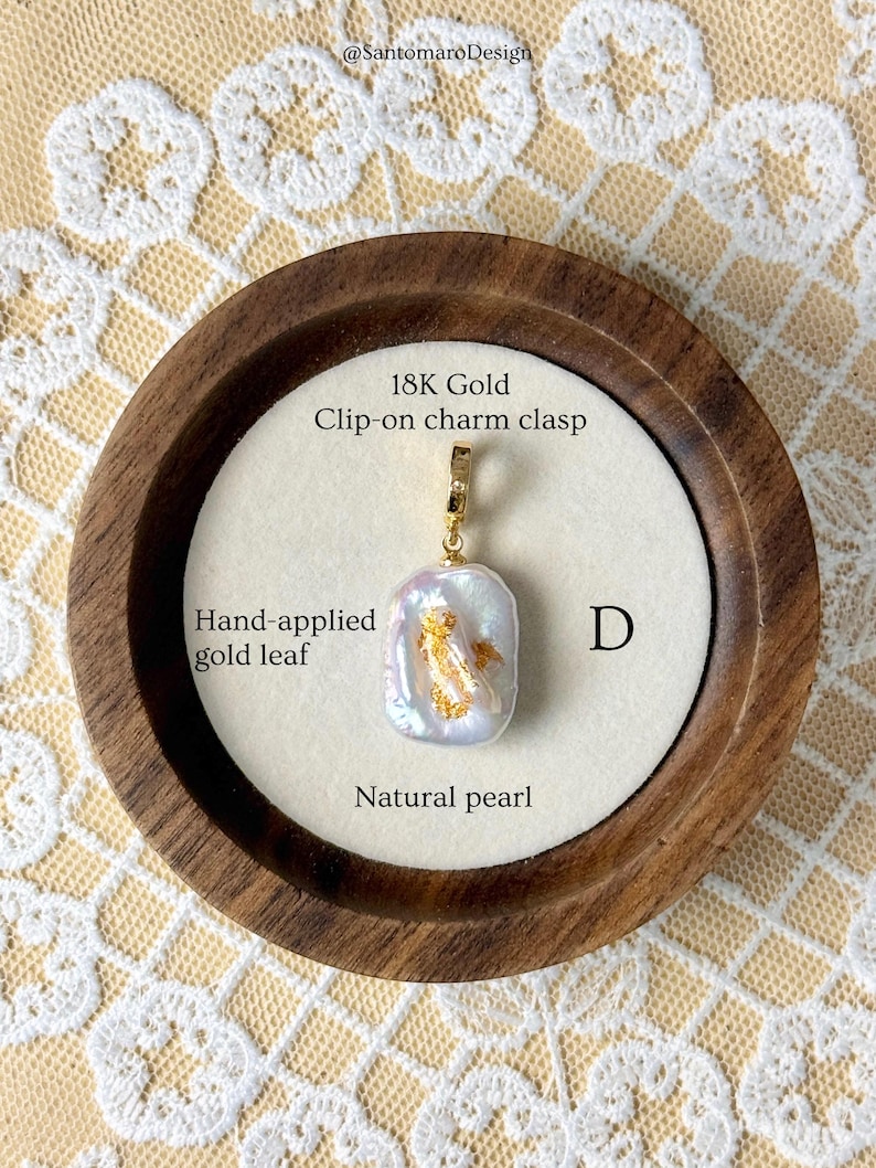 May include: A natural pearl pendant with hand-applied gold leaf, set in an 18K gold clip-on charm clasp. The pendant is displayed on a white surface within a wooden frame, with the text "Natural pearl" and "Hand-applied gold leaf".