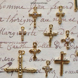 May include: A collection of gold-colored cross charms in various designs. Some crosses feature clear gemstone accents. The charms are displayed against a backdrop of handwritten script. The image is well-lit, highlighting the details of each charm.