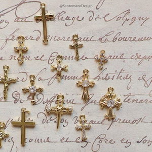 May include: A collection of gold-colored cross charms in various designs, some with clear gemstone accents. The charms are displayed against a backdrop of vintage script, with the designer's name, @SantomaroDesign, visible in the upper right corner.