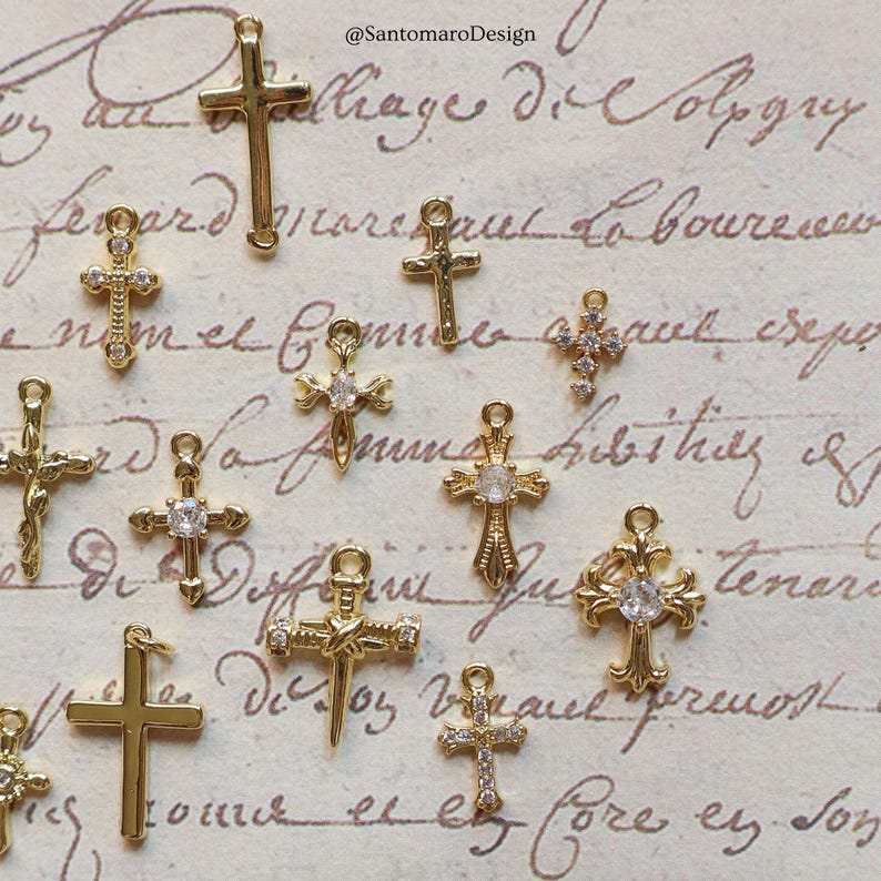 May include: A collection of gold-colored cross charms in various designs, some with clear gemstone accents. The charms are displayed against a backdrop of vintage script, with the designer's name, @SantomaroDesign, visible in the upper right corner.