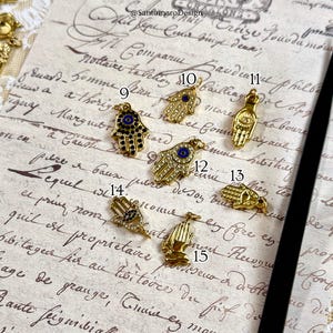 May include: Several gold-coloured Hamsa hand charms, some with black enamel, clear crystals, and blue detailing. The charms are presented on a vintage paper background, with the text "@SantomaroDesign" visible.