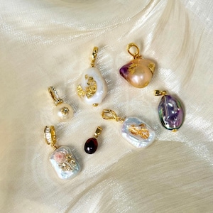 May include: Several gold pendants with pearls and gemstones are displayed. Some pendants have irregular white pearls with gold leaf, others have colourful gemstones. The pendants are presented on a cream-coloured, textured fabric. Text: @SantomaroDesign.