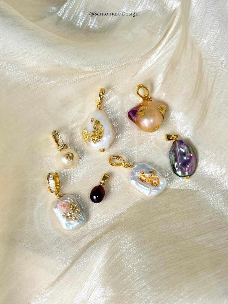 May include: Several gold pendants with pearls and gemstones are displayed. Some pendants have irregular white pearls with gold leaf, others have colourful gemstones. The pendants are presented on a cream-coloured, textured fabric. Text: @SantomaroDesign.