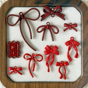 以下が含まれることがあります： 木製のトレイ内の白い表面に配置された、赤いリボン型のチャームと額入りのバラのデザインのコレクション。チャームは、リボンのようなデザインやクラシックなリボンの形など、さまざまなスタイルがあります。「@SantomaroDesign」の文字が見えます。