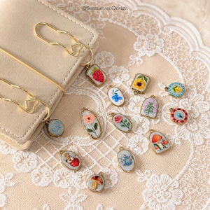 Può includere: Una collezione di ciondoli in miniatura dipinti a mano con cornici in metallo color oro. I ciondoli presentano vari disegni, tra cui fiori, frutta e funghi. Alcuni ciondoli sono attaccati a fermagli dorati. Lo sfondo è un pizzo color crema.