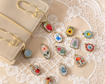 Könnte beinhalten: Eine Sammlung von handbemalten Miniatur-Anhängern mit goldfarbenen Metallrahmen. Die Anhänger zeigen verschiedene Designs, darunter Blumen, Früchte und Pilze. Einige Anhänger sind an goldfarbenen Büroklammern befestigt. Der Hintergrund ist eine cremefarbene Spitze.