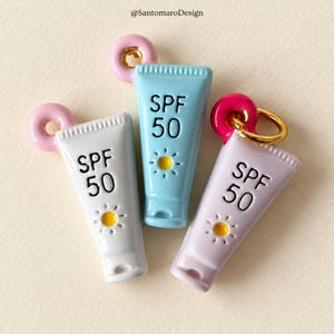 Può includere: Tre ciondoli in smalto a forma di tubetti di crema solare bianchi, azzurri e color lavanda. Ogni tubetto riporta la scritta nera SPF 50 e un'icona del sole gialla. I ciondoli hanno anelli rosa o magenta e un anellino dorato.