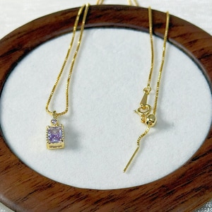 May include: A gold-colored necklace with a square, light purple pendant. The pendant is set in a gold frame and has a small, round purple stone above it. The necklace is displayed on a white surface within a wooden frame.