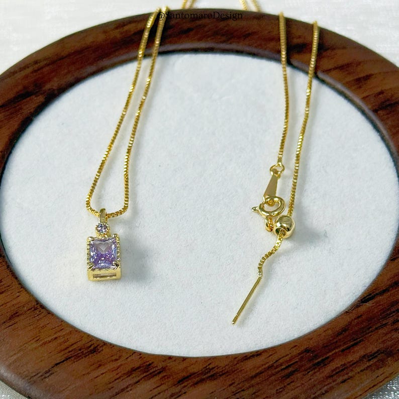 May include: A gold-colored necklace with a square, light purple pendant. The pendant is set in a gold frame and has a small, round purple stone above it. The necklace is displayed on a white surface within a wooden frame.