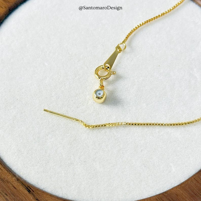 May include: Close-up of a gold-colored necklace with a delicate chain and a small, round charm. The charm has a light blue center with a white border. The necklace is on a white surface. The text "@SantomaroDesign" is in the upper left corner.