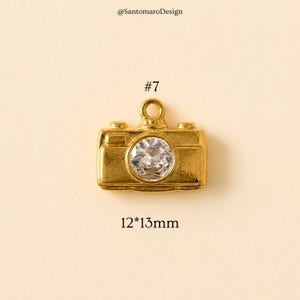 May include: A gold-colored camera charm with a clear, round gem in the center. The charm is approximately 0.47 x 0.51 inches in size, with the number "7" and the text "@SantamaroDesign" visible.