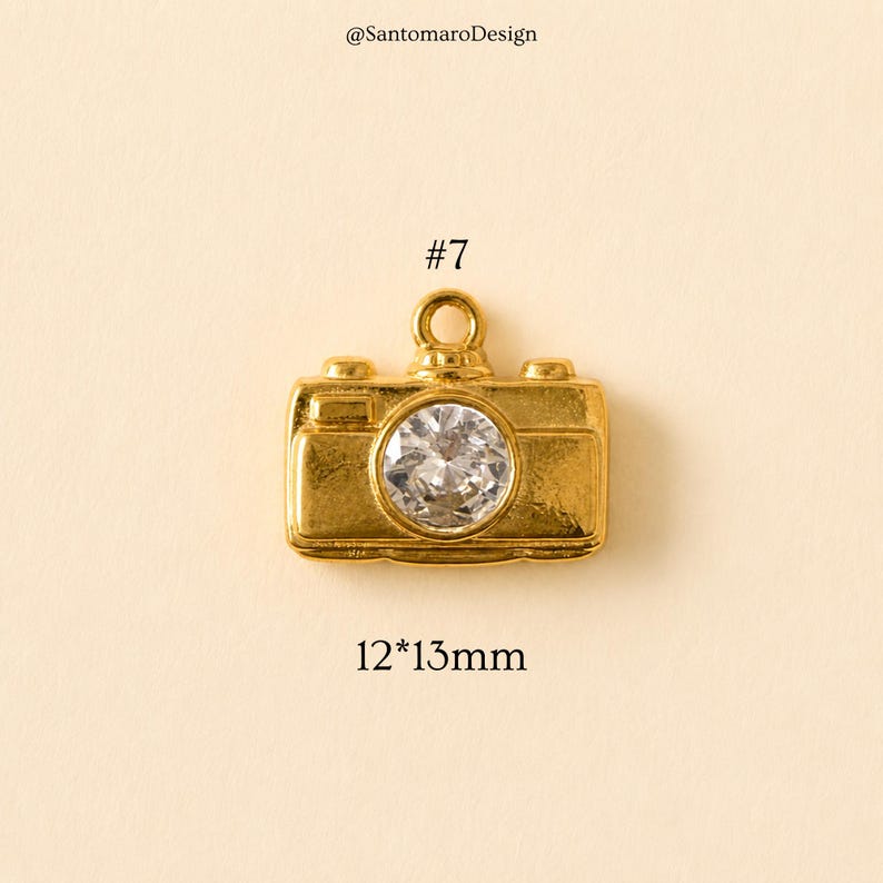 May include: A gold-colored camera charm with a clear, round gem in the center. The charm is approximately 0.47 x 0.51 inches in size, with the number "7" and the text "@SantamaroDesign" visible.
