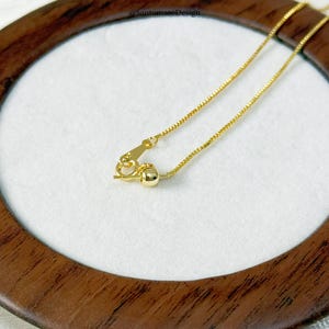 May include: Close-up of a gold-colored necklace with a delicate chain and a small clasp. The necklace is displayed on a white surface within a wooden frame. The chain has a box link design.