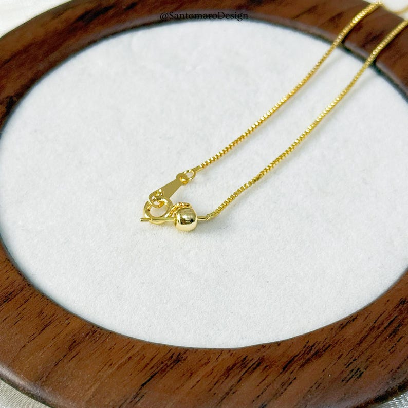 May include: Close-up of a gold-colored necklace with a delicate chain and a small clasp. The necklace is displayed on a white surface within a wooden frame. The chain has a box link design.