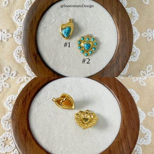 Peut inclure: Deux pendentifs dor&eacute;s en forme de c&oelig;ur avec des accents turquoise et un m&eacute;daillon dor&eacute; en forme de c&oelig;ur sont pr&eacute;sent&eacute;s dans une bo&icirc;te en bois. Les pendentifs sont &eacute;tiquet&eacute;s #1 et #2. Le texte "@SantomaroDesign" est visible.