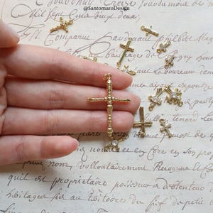 May include: A collection of gold-colored cross charms, some adorned with small, clear crystals. The crosses vary in design, with one larger, ornate cross held in a hand. The background features handwritten script.