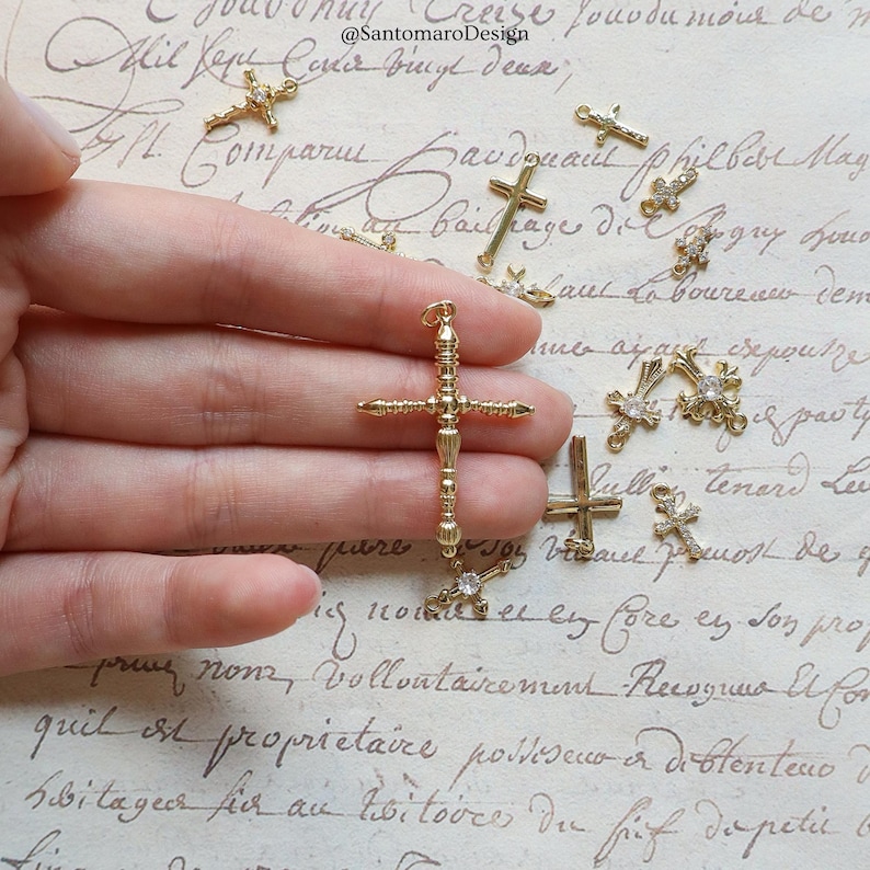 May include: A collection of gold-colored cross charms, some adorned with small, clear crystals. The crosses vary in design, with one larger, ornate cross held in a hand. The background features handwritten script.
