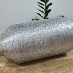 Puede incluir: Una bobina de hilo metálico plateado. El hilo está enrollado firmemente alrededor de una bobina de cartón con una etiqueta roja y blanca.