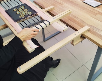 Könnte beinhalten: Ein hölzerner Stickrahmen mit einem rosa Stoff, der darüber gespannt ist. Eine Person arbeitet an einem Stickprojekt mit einem schwarzen und goldenen Design. Der Rahmen ist an einem Holzständer befestigt.