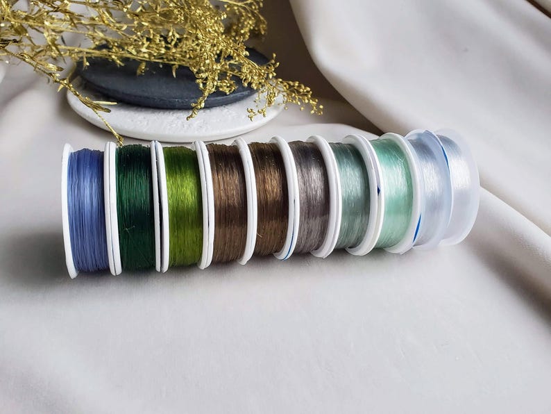 May include: Assortment of eight spools of elastic cord in various colours, including blue, green, brown, grey, and clear. The spools are arranged in a row, with the blue spool on the left and the clear spool on the right.
