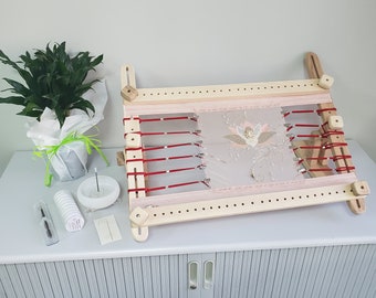Può includere: Un telaio da ricamo in legno con cinghie rosse e un tessuto rosa teso sopra. Il tessuto è ricamato con un motivo floreale. Il telaio è su un tavolo con una pianta, un piatto bianco e un rotolo di filo.