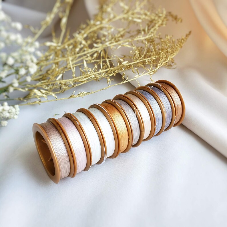 May include: A collection of spools of thread in various shades of white, beige, and grey. The spools are arranged in a stack, with the gold coloured plastic bobbins visible.