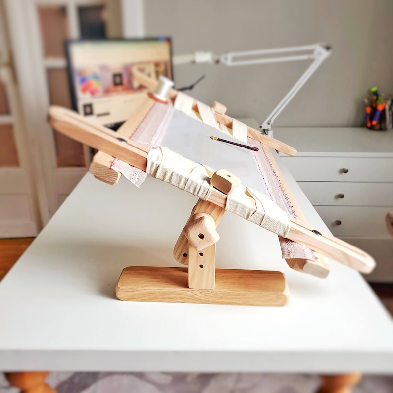 Op de afbeelding: Een houten borduurstandaard met een wit doek eroverheen gespannen. De standaard is verstelbaar en heeft een voet die het mogelijk maakt om hem in verschillende hoeken te positioneren.