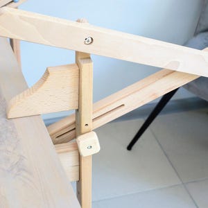 May include: A wooden table with a folding mechanism. The table is made of light wood and has a natural finish. The folding mechanism is made of metal and is attached to the underside of the table.