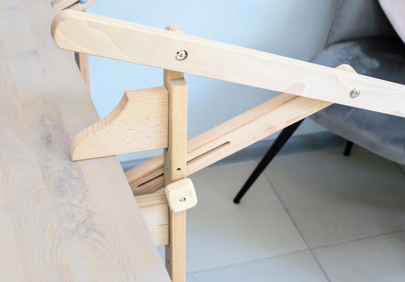 May include: A wooden table with a folding mechanism. The table is made of light wood and has a natural finish. The folding mechanism is made of metal and is attached to the underside of the table.