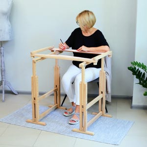 Puede incluir: Un bastidor de bordado de madera con altura ajustable, con una persona sentada y trabajando en un trozo de tela. El bastidor tiene un acabado en madera clara y está diseñado para un bordado cómodo.