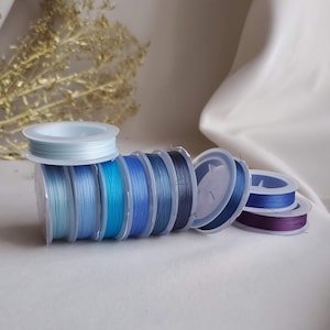 Puede incluir: Una colección de carretes de cordón elástico azul y azul claro, enrollados en carretes de plástico transparente. Los carretes están dispuestos en una pila, con el cordón azul claro en la parte superior.