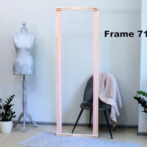 Puede incluir: Un marco de madera de color rosa claro para tejer, que mide 71 pulgadas de largo. El marco está sobre una alfombra gris frente a una pared blanca.
