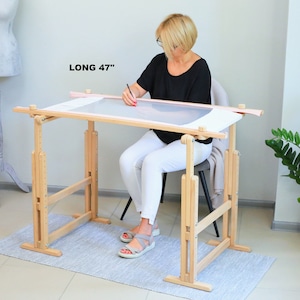 May include: A wooden embroidery stand with adjustable height, supporting a white canvas. The stand is 47 inches long and is being used by a person sitting in a chair.