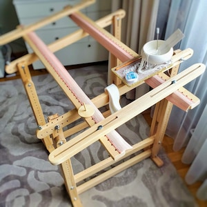 May include: A wooden embroidery stand with a light pink fabric stretched across it. The stand has adjustable wooden arms and a small tray holding embroidery supplies. A white bowl with thread and a needle are also visible.
