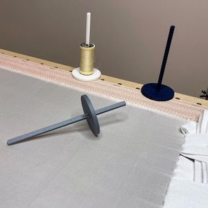 Peut inclure: Un métier à broder avec un tissu rose clair tendu dessus. Un enfile-aiguille gris, une bobine de fil doré et un support de fil bleu foncé sont sur le tissu. Le cadre en bois est visible.