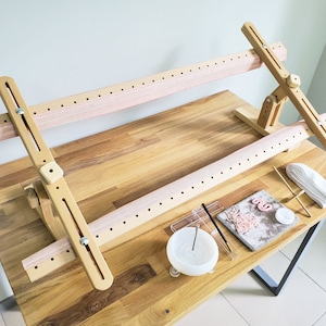 May include: A wooden embroidery hoop stand with two adjustable hoops, a pink fabric band, and a wooden base. The stand is on a wooden table with embroidery supplies, including a white fabric, a small wooden stand, a white cotton pad, and a small clear plastic container with a needle.