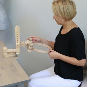 May include: A wooden embroidery hoop stand with a clamp to attach to a table. The stand has adjustable arms to hold the hoop at different angles. A person is shown using the stand to hold a hoop.