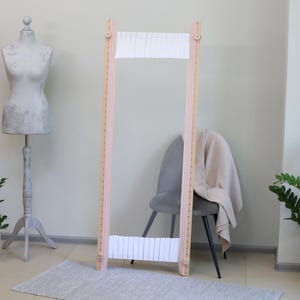 May include: A wooden loom with white warp threads, standing on a light grey rug in a room with a grey chair and a plant. The loom is pink and has a natural wood finish.