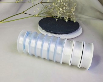 May include: A stack of eight clear plastic spools of white elastic thread. The spools are arranged in a circle, with the largest spool at the bottom and the smallest spool at the top.