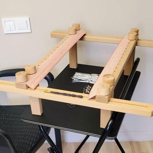 May include: A wooden embroidery frame with a pink fabric stretched across it. The frame is sitting on a black table with a small wooden tool and white string.