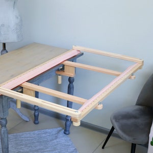 以下が含まれることがあります： ピンクの布が張られた木製の手芸用枠。枠はクランプでテーブルに取り付けられています。