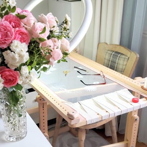 May include: A wooden embroidery hoop stand with a white fabric stretched across the hoop. The stand has a wooden frame and legs. A spool of white thread is on the stand. A bouquet of pink and white roses is in a clear glass vase on a white table.