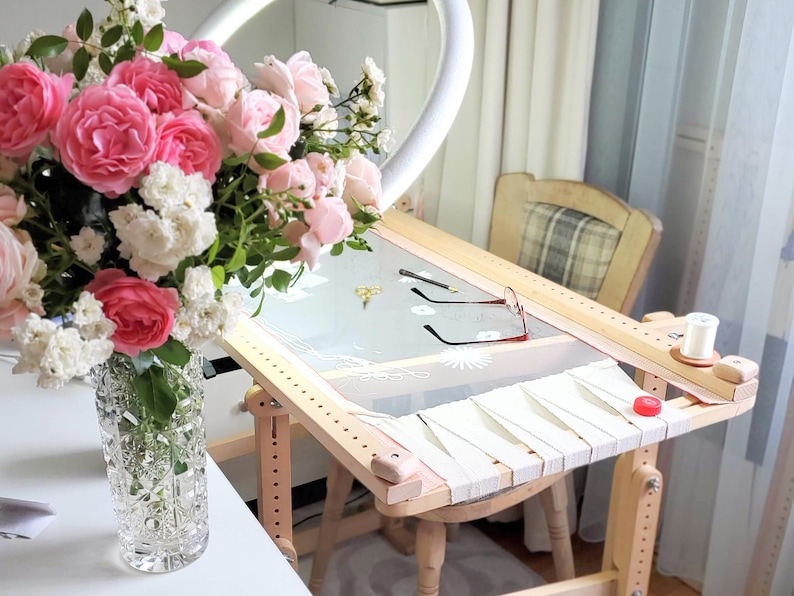 May include: A wooden embroidery hoop stand with a white fabric stretched across the hoop. The stand has a wooden frame and legs. A spool of white thread is on the stand. A bouquet of pink and white roses is in a clear glass vase on a white table.