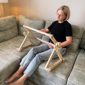 Puede incluir: Un soporte de bastidor de bordado de madera, ajustable en altura y ángulo. El soporte es de madera clara y tiene un marco rectangular para sujetar la tela. Diseñado para proyectos de bordado y punto de cruz a manos libres.