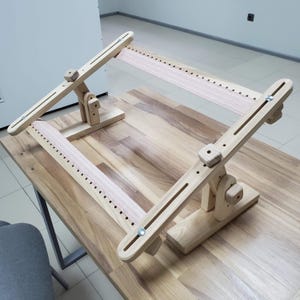 以下が含まれることがあります： 調節可能なアームと上部に張られたピンクの布が付いた木製刺繍スタンド。スタンドは、作業中の刺繍プロジェクトを保持するために設計されています。
