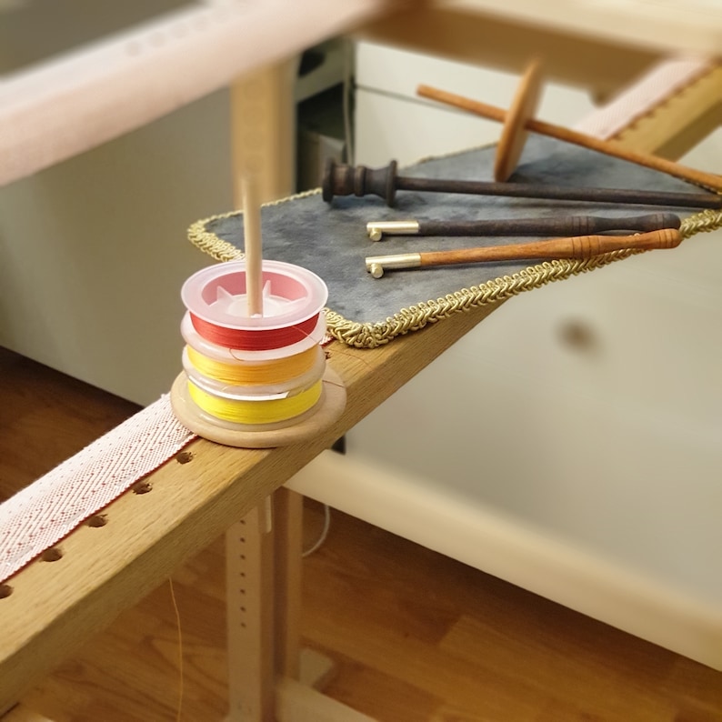 May include: A wooden embroidery stand with a spool holder containing three spools of thread in red, yellow, and orange. The stand has a small wooden dowel for holding the thread. There are also several wooden tools on a grey mat with gold trim.