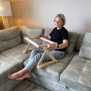 Puede incluir: Un soporte de bordado de madera con un soporte de tela rosa claro. El soporte es ajustable y tiene una base triangular. El soporte está diseñado para trabajos de aguja y se muestra en uso en un sofá gris.