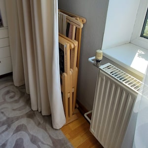 May include: A wooden folding loom for weaving, standing upright against a wall. The loom is made of light wood and has a natural finish. It is folded up and leaning against a white wall. The loom is in a room with a white radiator and a window.