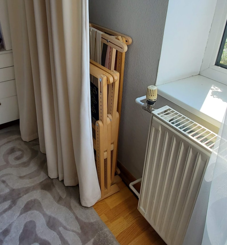 May include: A wooden folding loom for weaving, standing upright against a wall. The loom is made of light wood and has a natural finish. It is folded up and leaning against a white wall. The loom is in a room with a white radiator and a window.